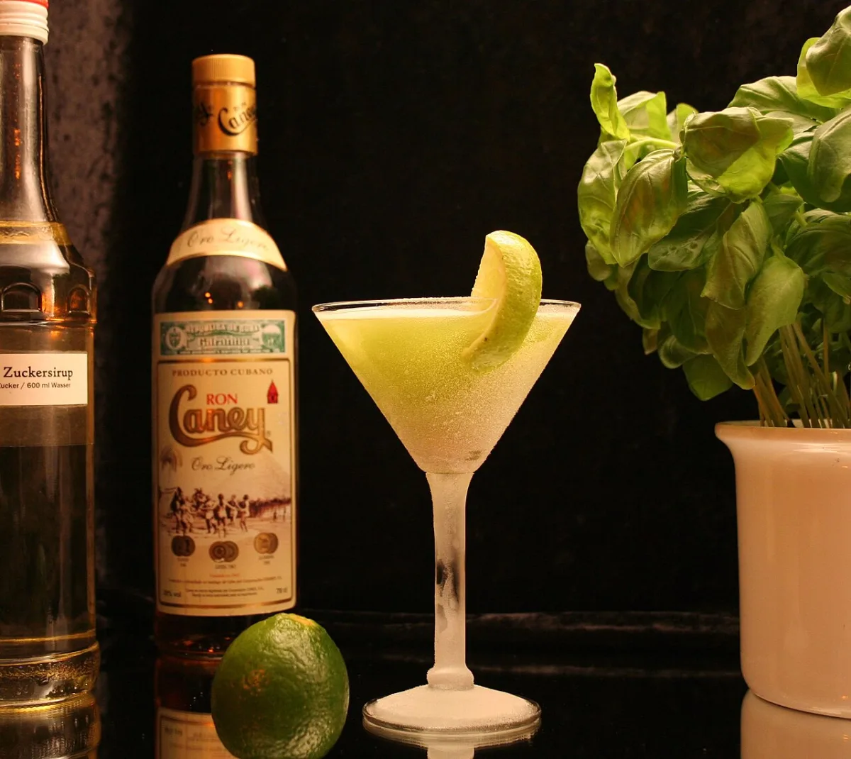 Daiquiri cocktail met limoen in een martiniglas naast een fles rum en verse basilicum