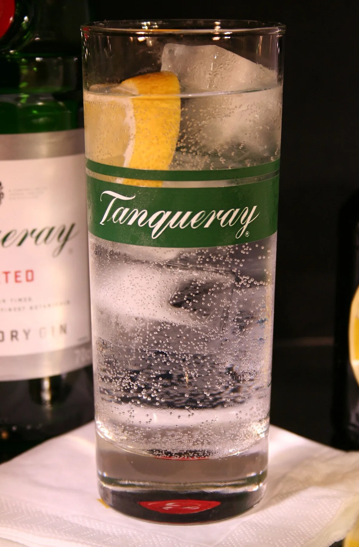 Glas gin-tonic met citroenschijfje en ijsblokjes, met een Tanqueray ginfles op de achtergrond