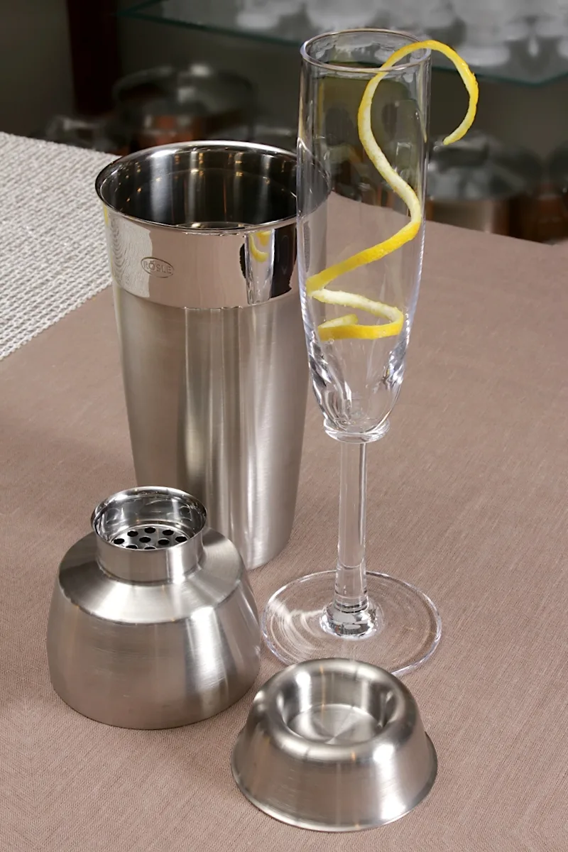 Roestvrijstalen cocktailshaker met onderdelen naast een champagneflûte met citroenschil