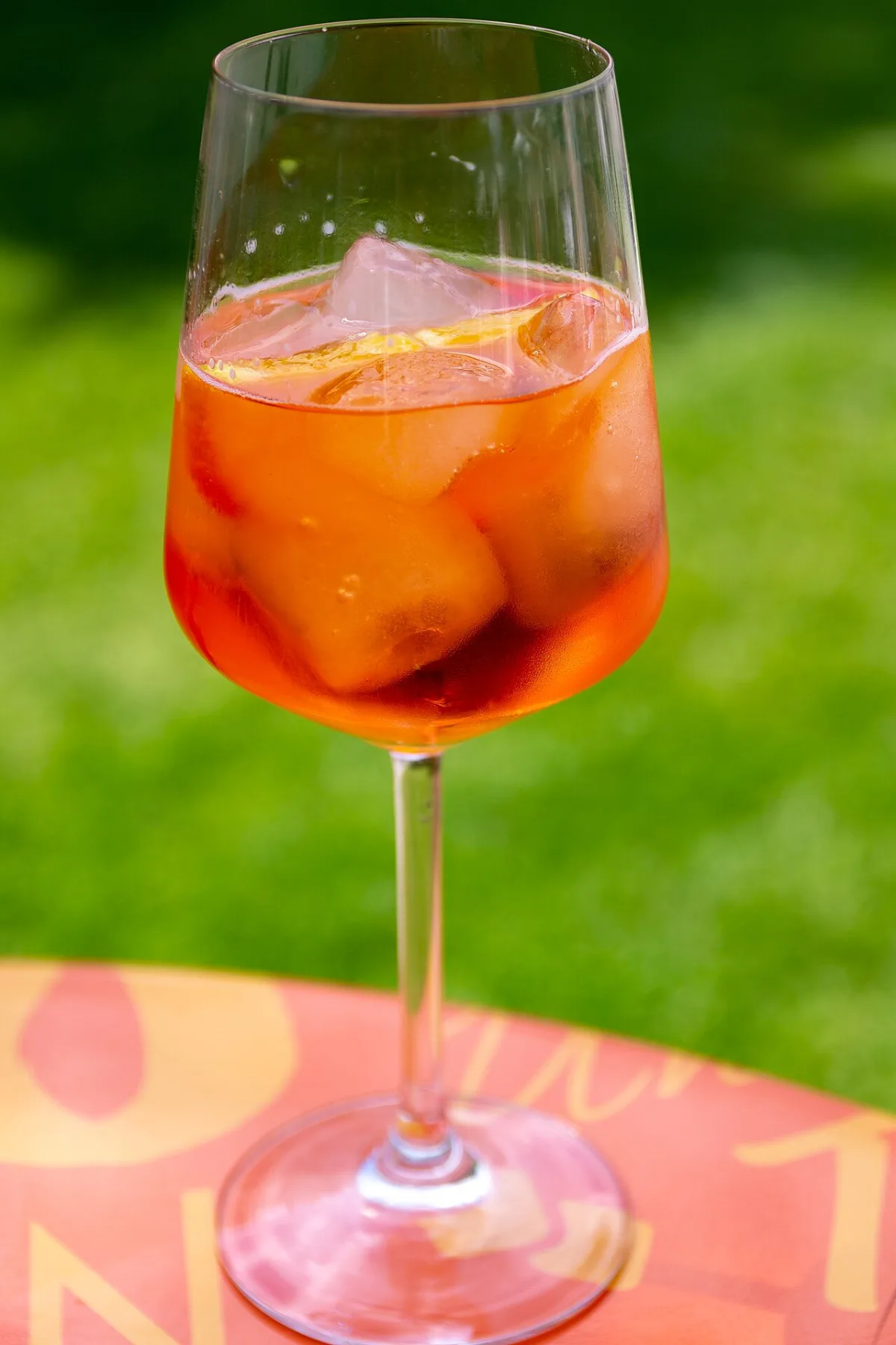 Glas Aperol Spritz met ijsblokjes en sinaasappelschijf buiten op een tafel met groen gras op de achtergrond